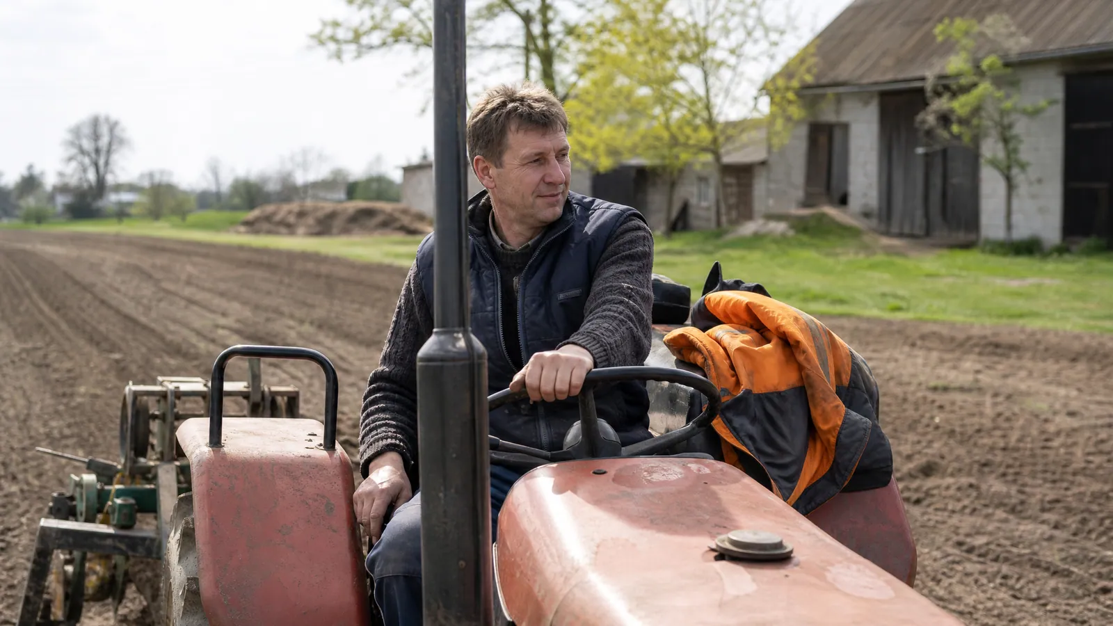 Rolnik w kabinie ciągnika pracuje na polu obok zabudowań gospodarstwa